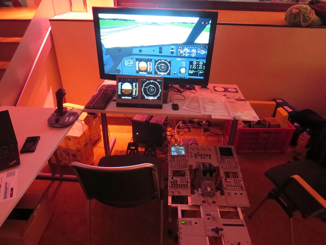 The setup consists of two tables arranged in an L shape, one behind it and one to the side where the side stick rests on. On the table in the back is a big monitor showing the simulator and a smaller one, showing the primary flight display and navigation display. In front of the monitors is a chair for the pilot flying, to the right of it is the DIY pedestal made out of many 3d printed panels with lots of buttons, knobs and switches. To the right of that is a second chair for a pilot monitoring. Behind the desk there are some boxes which were used to transport the sim as well as the PC running the simulation.